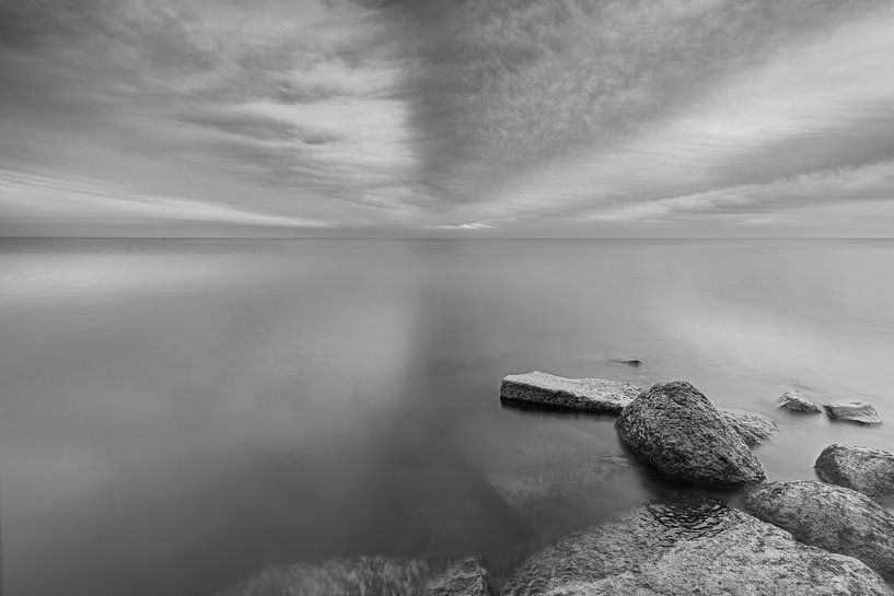 Black and white photo of threatening striped clouds over a calm Markermeer with stones as foreground by Bram Lubbers