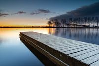 Der schneebedeckte Steg und die Baumreihe spiegeln sich bei Sonnenaufgang im ruhigen See