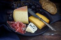 Eetbordje met diverse kaassoorten, luchtgedroogde ham, blauwe druiven en brood op donker rustiek hou