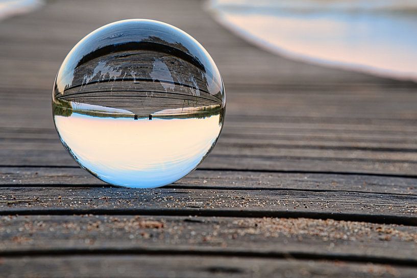 Glass ball on a footbridge reaching into a lake by Martin Köbsch
