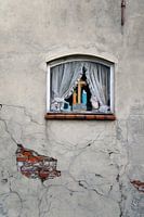 Verweerde muur met een raam met gordijnen en kruis met een Mariabeeld