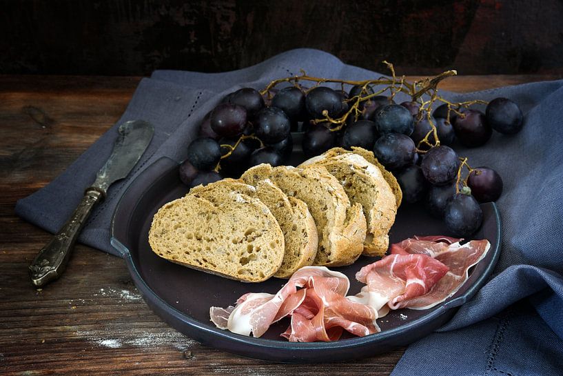 pain complet frais avec des raisins bleus et du jambon de Prosciutto sur une table en bois sombre et par Maren Winter