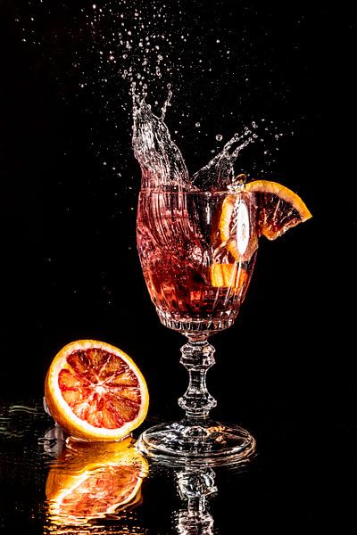 Blood orange piece falls into a glass with water mirrored on black background by Hans-Jürgen Janda