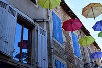 Parapluies colorés suspendus dans la rue, reflets dans les fenêtres, volets en bois à côté.